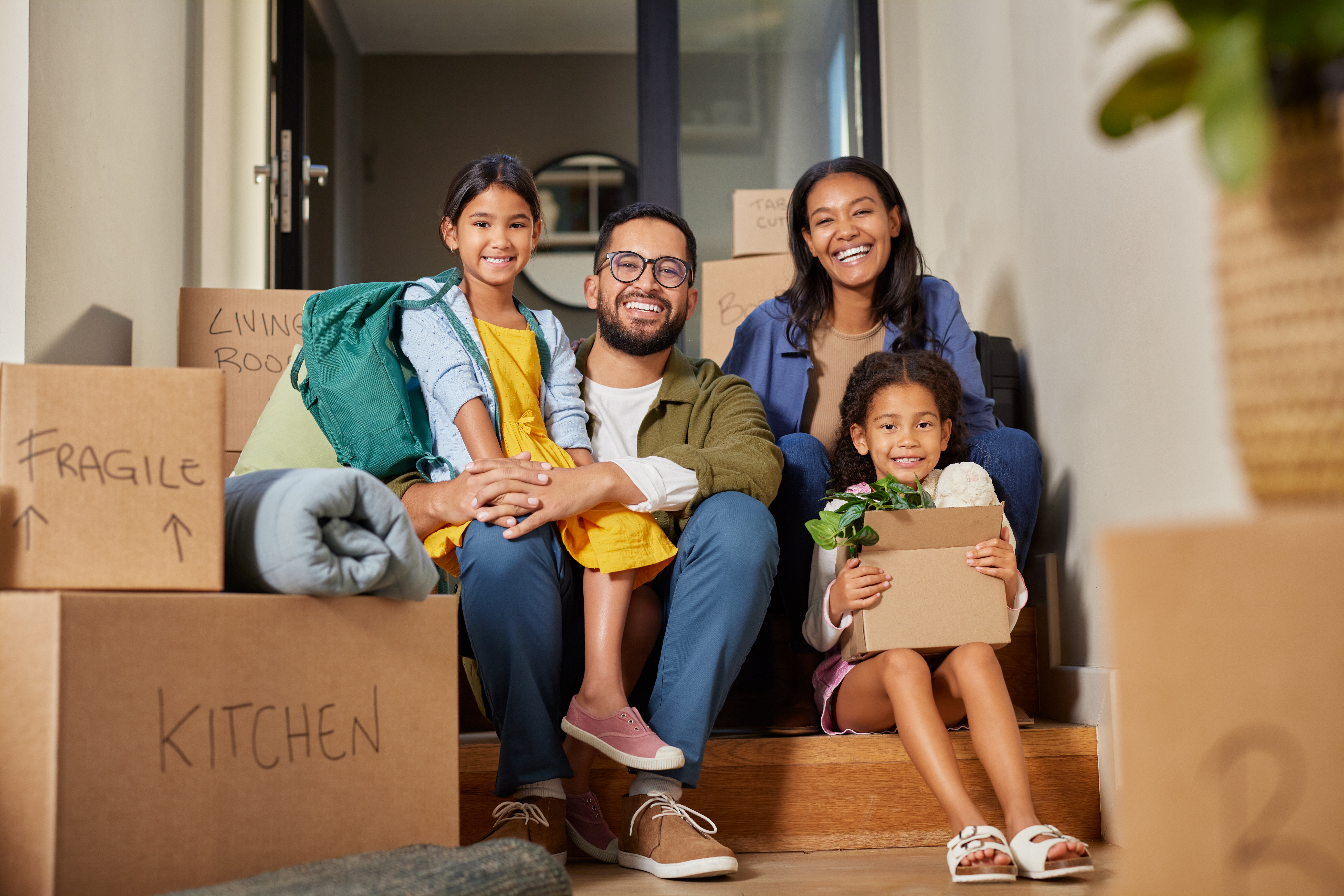 Two parents and their daughters sitting on the stairs to their new house, surrounded by moving boxes.