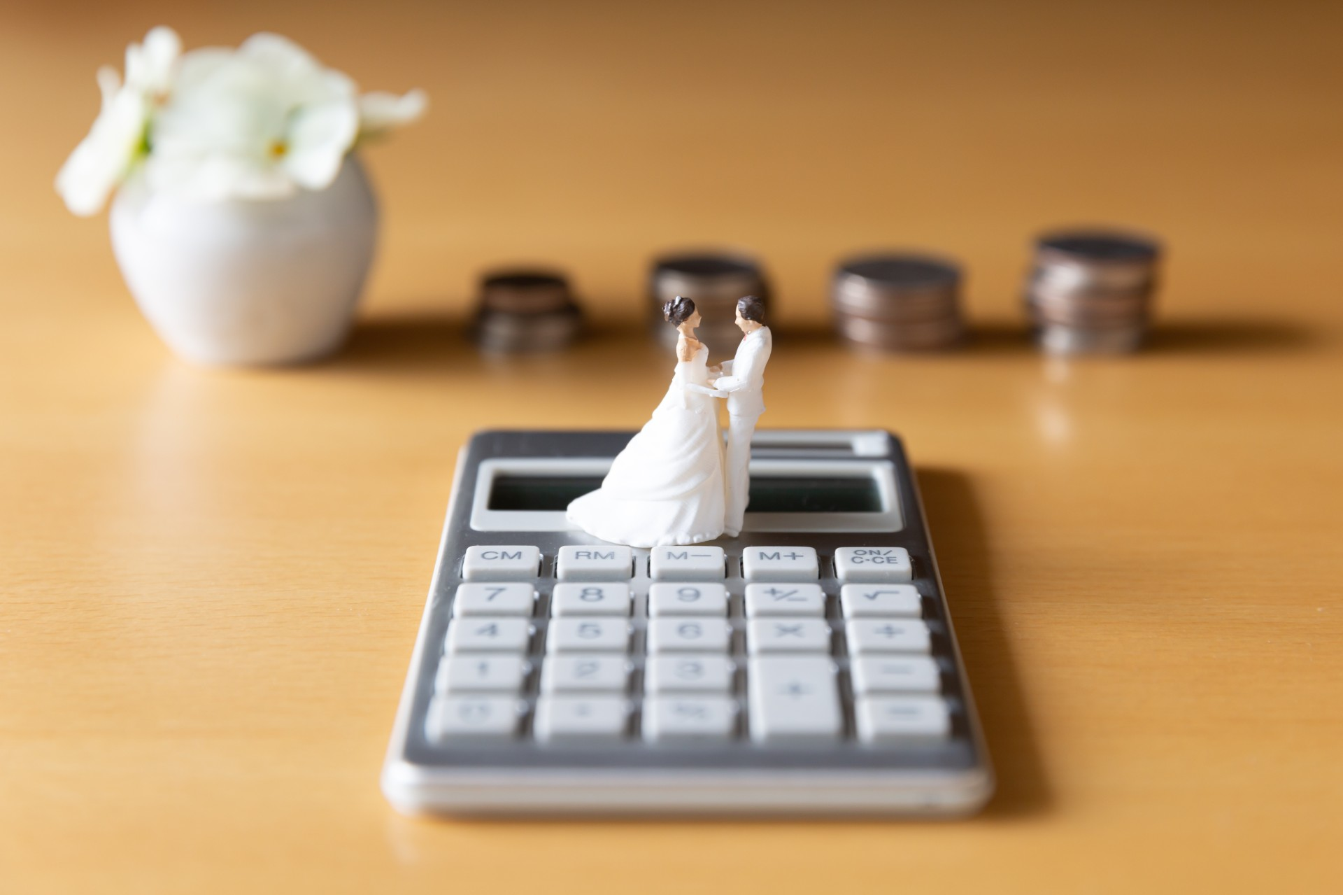 A wedding cake topper, a wife and husband, on top of a calculator. Behind them are white flowers and quarters stacked.